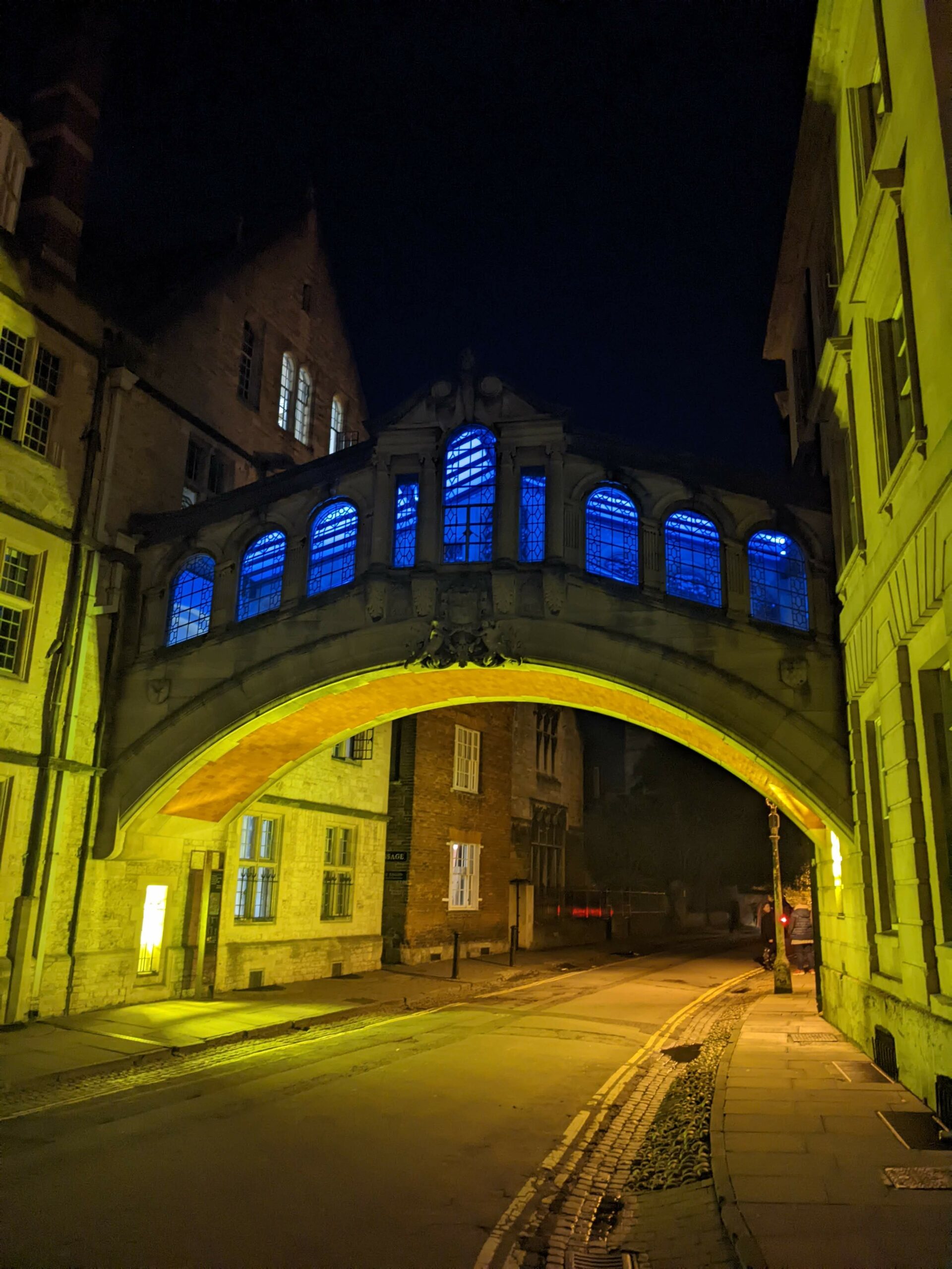 Night-time photograph of stone bridge illuminated in yellow with blue lights shining from windows