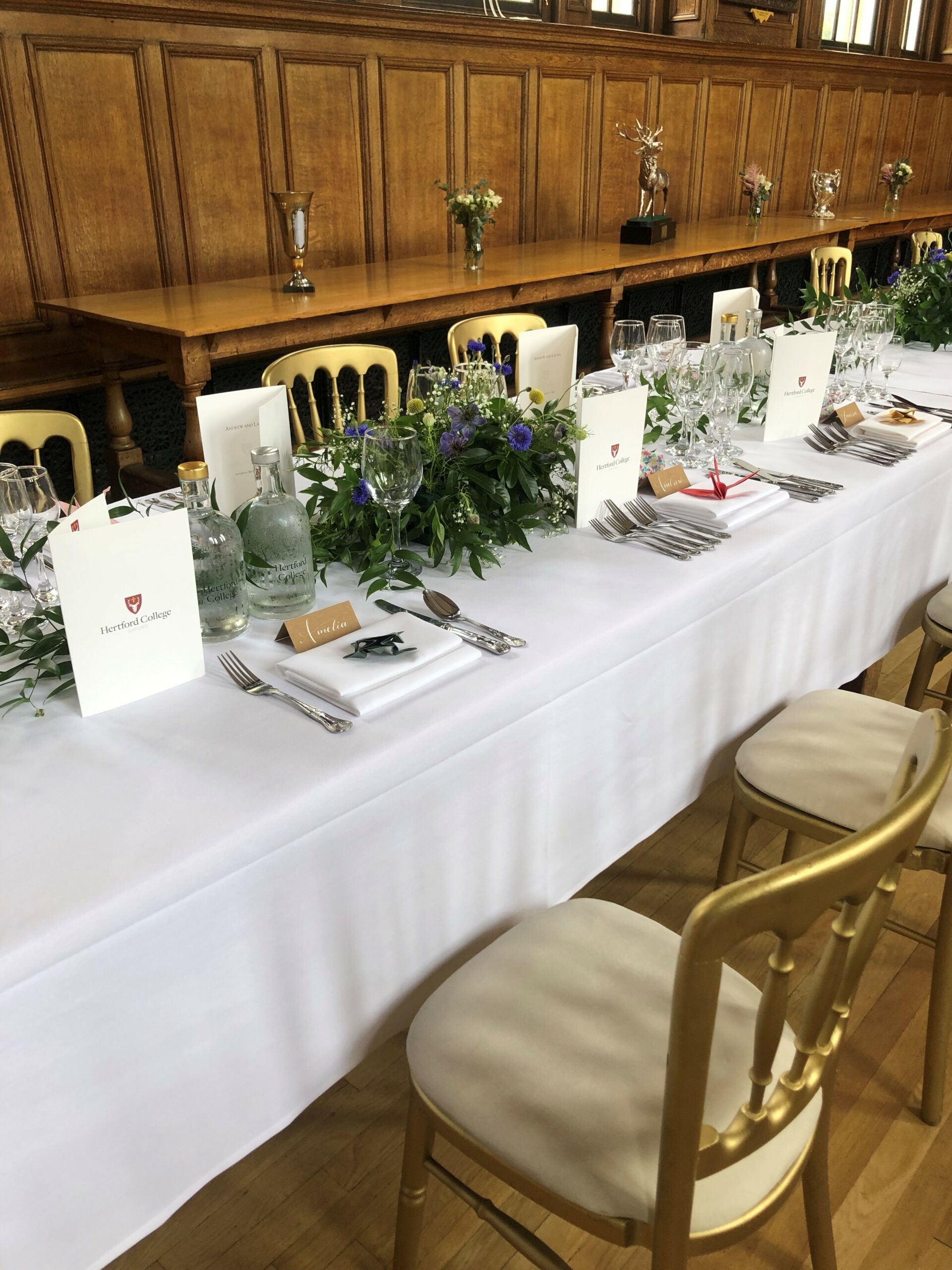 A long table with white table cloth, flower arrangements and gold chairs